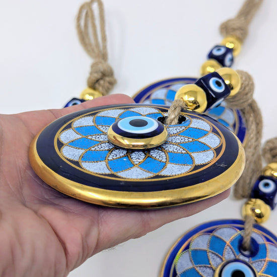 A hand holding a round blue and gold evil eye ornament with a tassel, with two more similar ornaments visible in the background against a plain white backdrop.