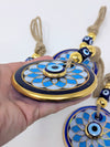 A hand holding a round blue and gold evil eye ornament with a tassel, with two more similar ornaments visible in the background against a plain white backdrop.