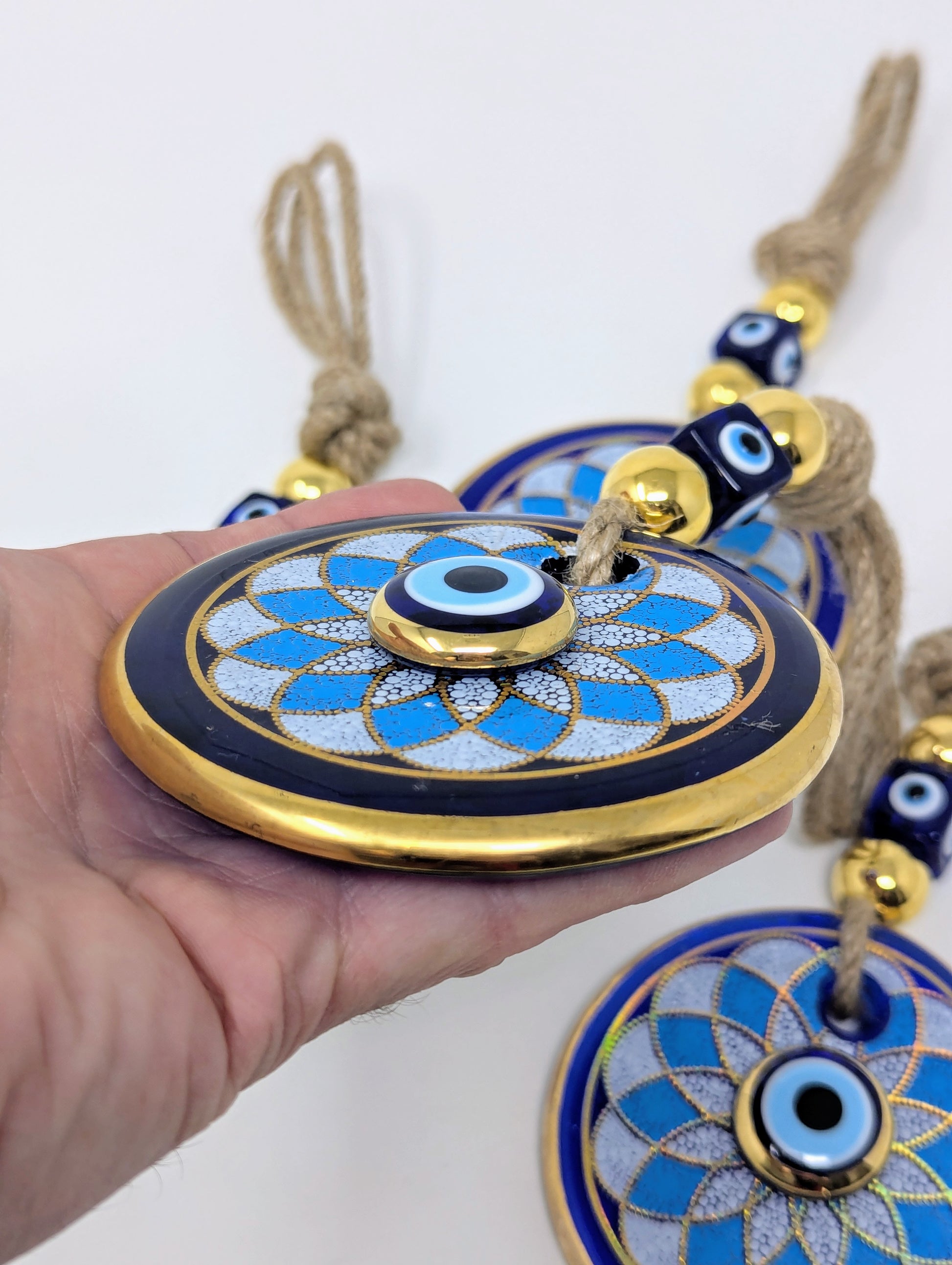 A hand holding a round blue and gold evil eye ornament with a tassel, with two more similar ornaments visible in the background against a plain white backdrop.