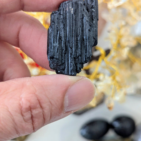A hand holding a small, dark, vertically textured mineral specimen against a softly blurred background with warm decorative elements.