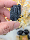A hand holding a small, dark, vertically textured mineral specimen against a softly blurred background with warm decorative elements.