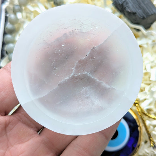 A hand holding a round, translucent white stone or crystal piece with natural cloudy veining, set against a background of shredded paper and small decorative items.