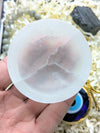 A hand holding a round, translucent white stone or crystal piece with natural cloudy veining, set against a background of shredded paper and small decorative items.