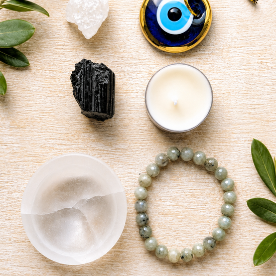 A flat-lay of a spiritual protection bundle on a light wood background, featuring a blue and gold evil eye pendant, a white salt shield, a black crystal, a green jade bracelet, a white candle, and crystals, with text reading 'PROTECTION BUNDLE' at the bottom.