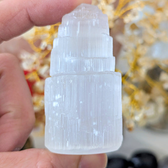 A hand holding a small, tiered white crystal tower with a ribbed cylindrical base, set against a blurred background of gold decorative branches.