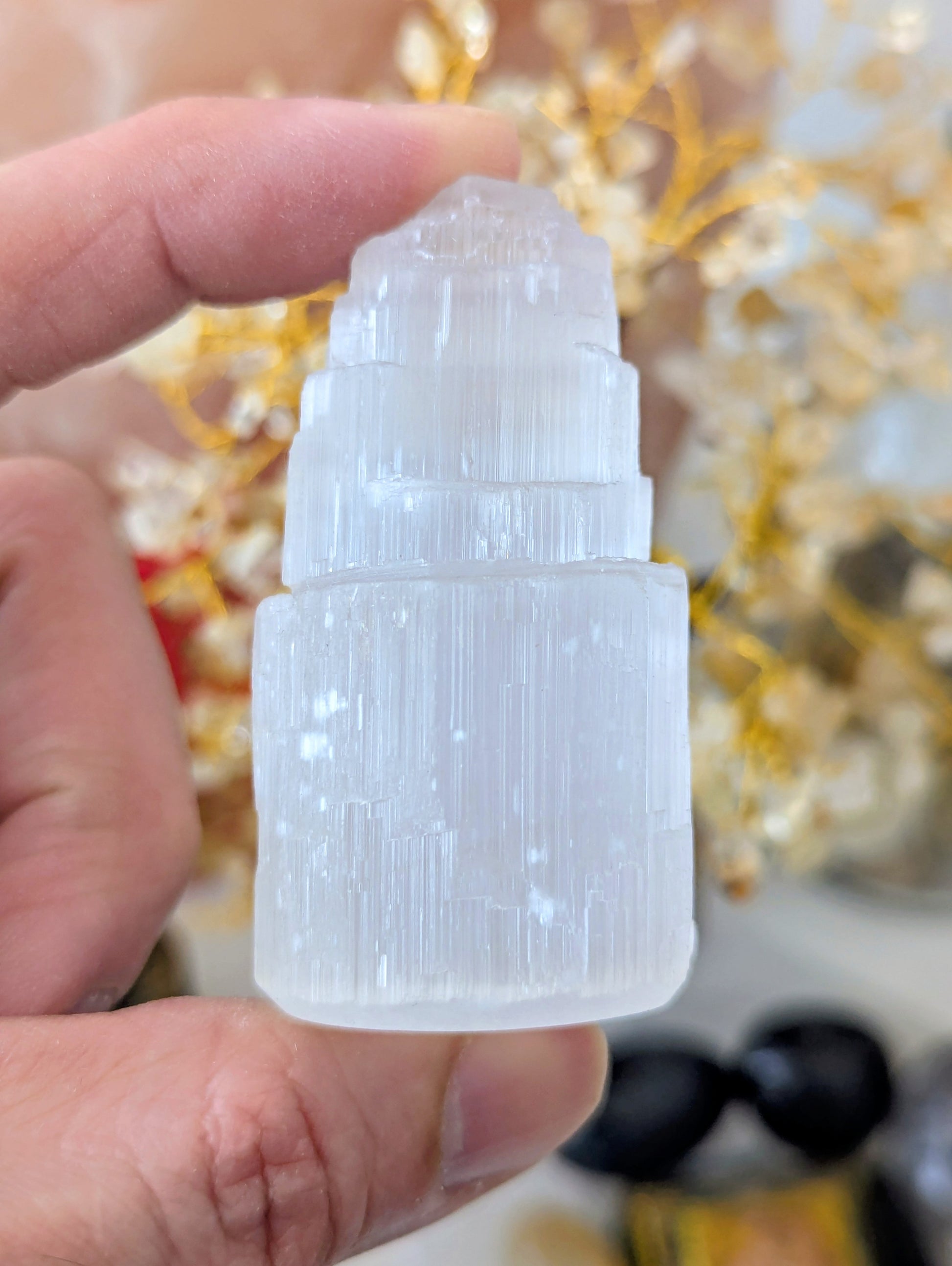 A hand holding a small, tiered white crystal tower with a ribbed cylindrical base, set against a blurred background of gold decorative branches.