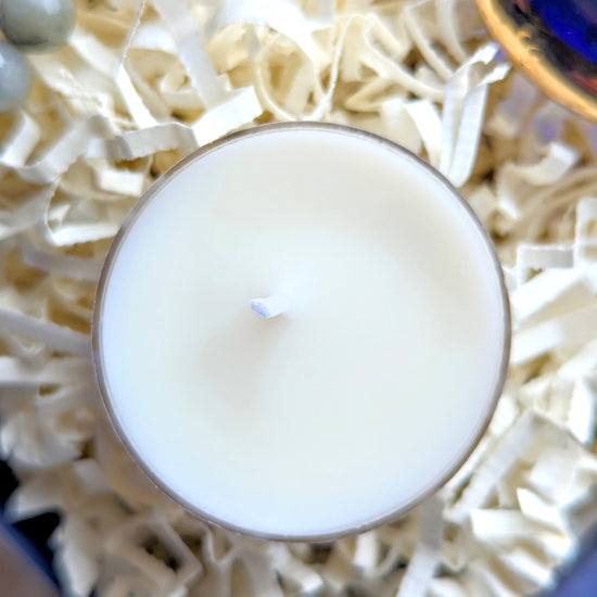 A top-down close-up of a white wax candle in a clear glass container, resting on cream-colored shredded paper filler.
