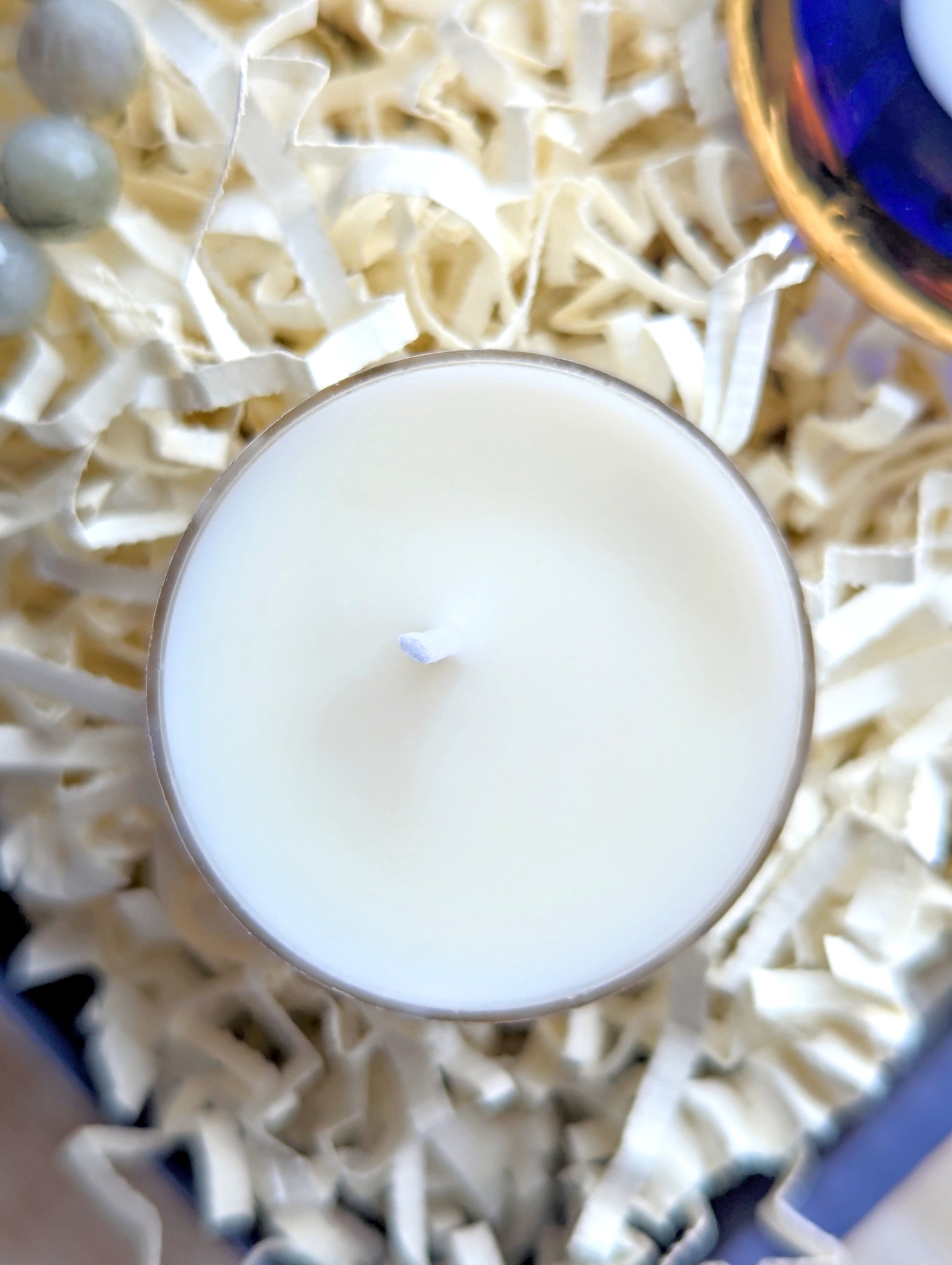 A top-down close-up of a white wax candle in a clear glass container, resting on cream-colored shredded paper filler.